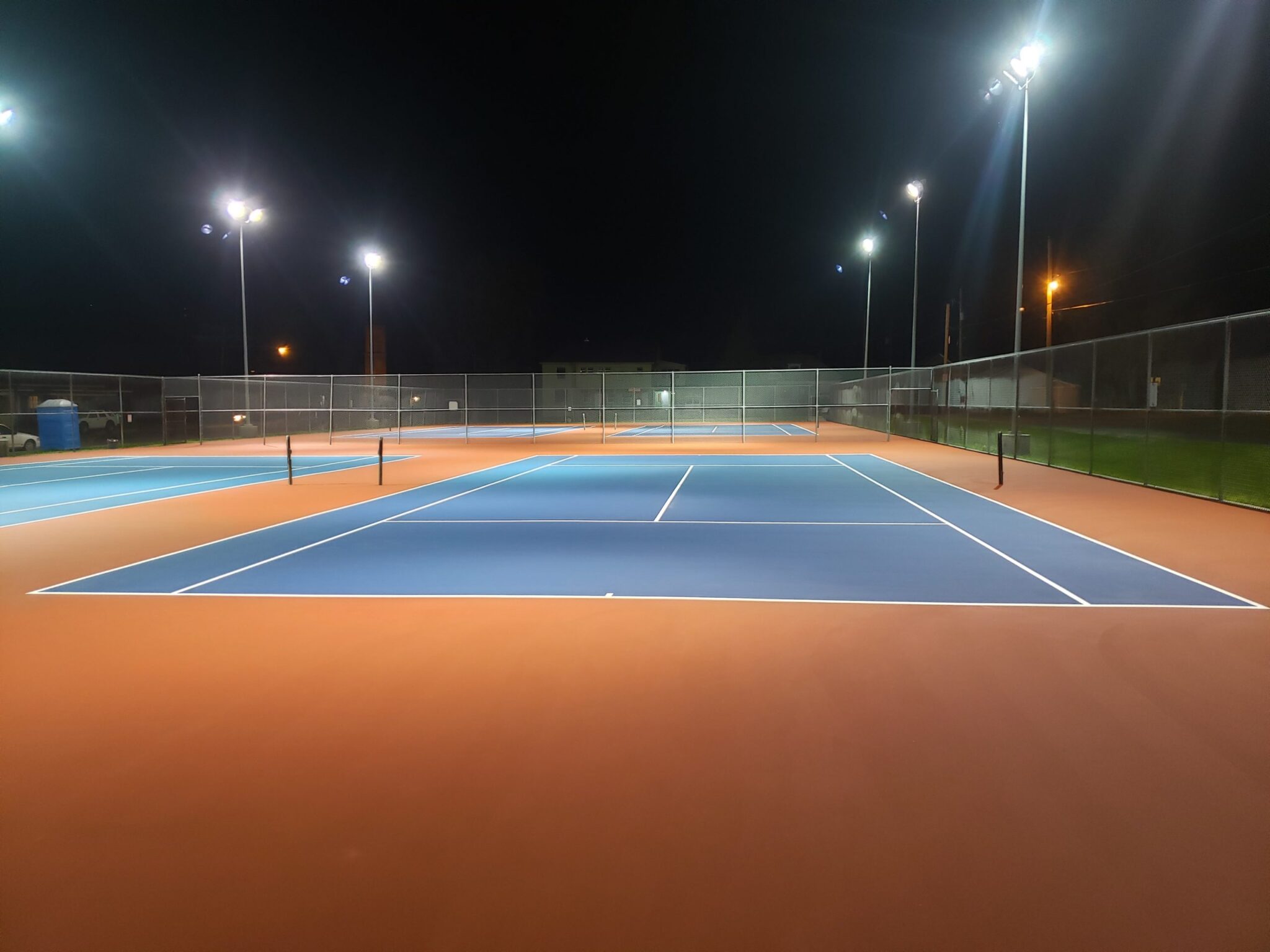 A new tennis court complex under lights with fencing that includes orange, blue, and white colors.
