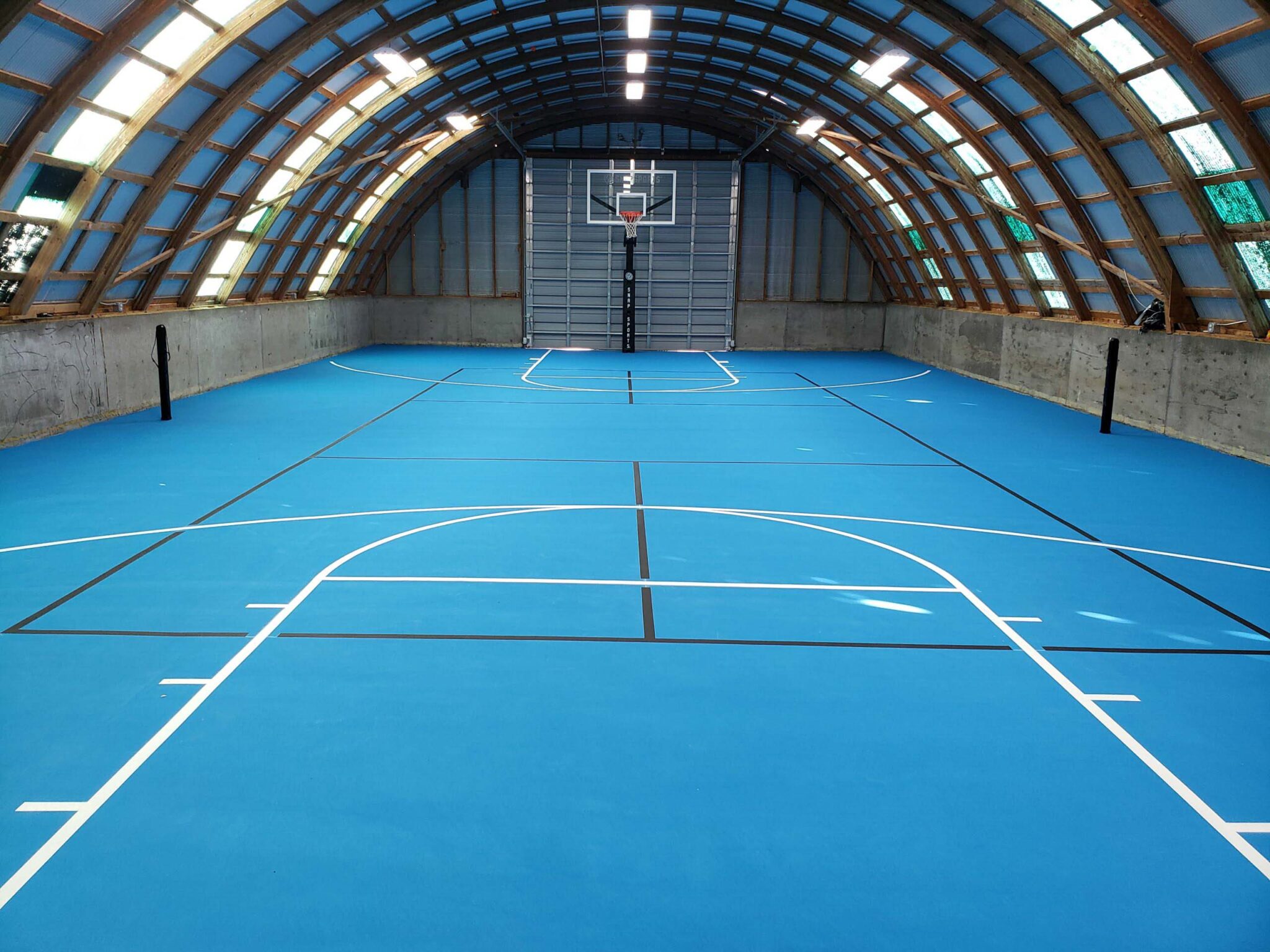 A basketball court including lines for pickleball where the court is blue, the basketball lines are white, and the pickleball lines are black. The court is enclosed in a domed garage.